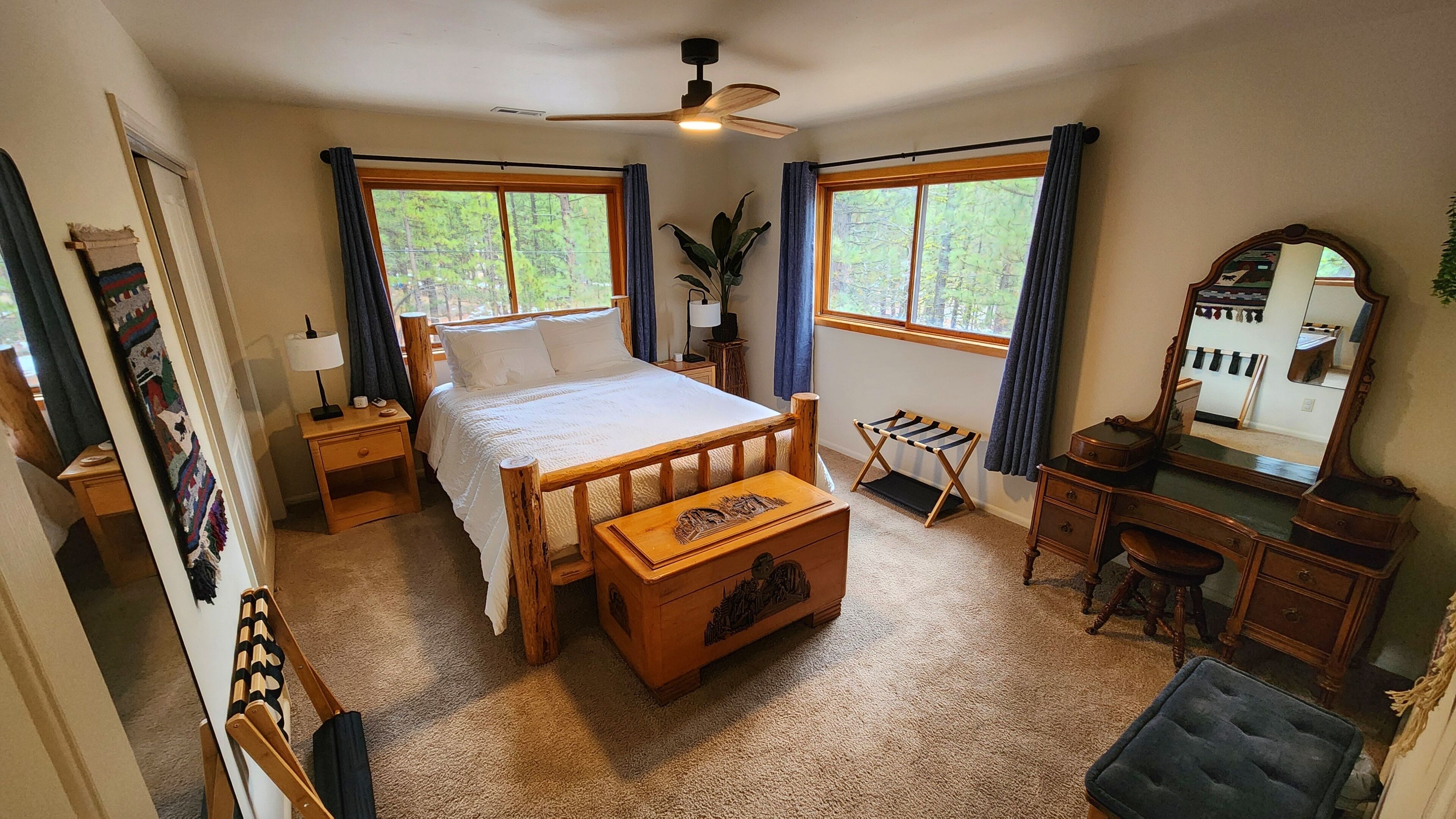 Un dormitorio con cama, mesitas de noche, cómoda, espejo y escritorio. Hay ventanas con cortinas, un ventilador de techo y suelo alfombrado.