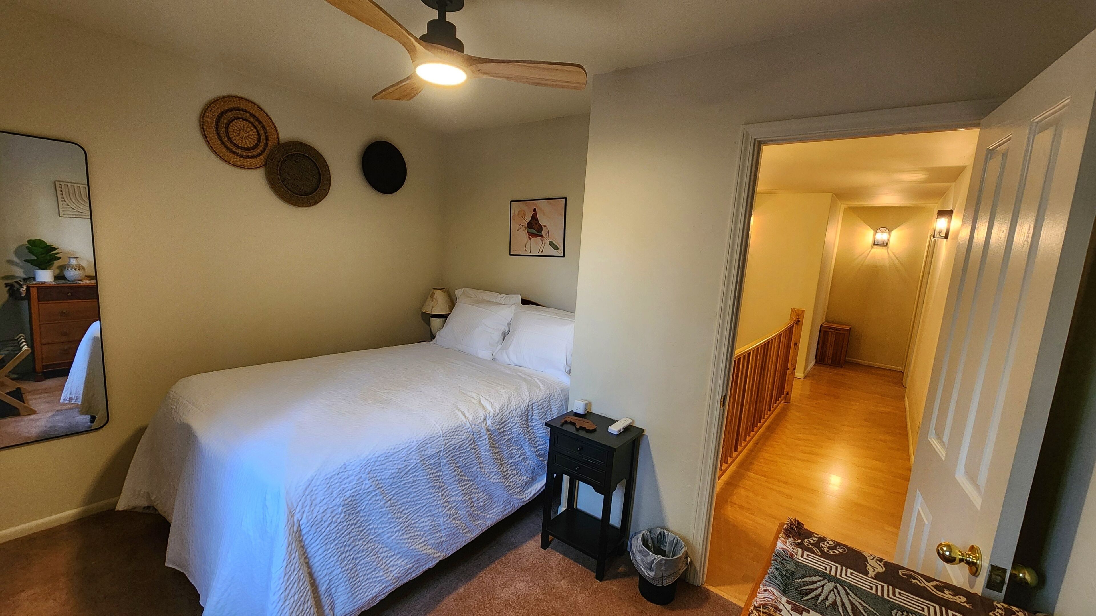 A bedroom with a bed, a ceiling fan, a nightstand, a trash can, and a door leading to a hallway.