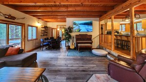 A spacious living area with a wooden ceiling, a dining table with chairs, a piano, and a large TV screen.