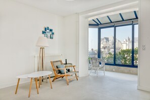 A bright room with a white table, a chair, a floor lamp, and a view of buildings through large windows.
