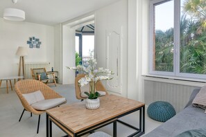 A room with a wooden table, wicker chairs, a sofa, and a window with a view of palm trees.