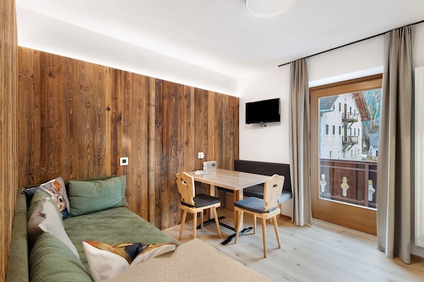 A room with a wooden wall, a sofa, a table with chairs, a TV, and a window with curtains.