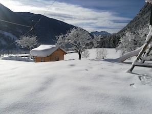 A wooden cabin with a snow-covered roof, surrounded by snow-covered trees and mountains in the background.