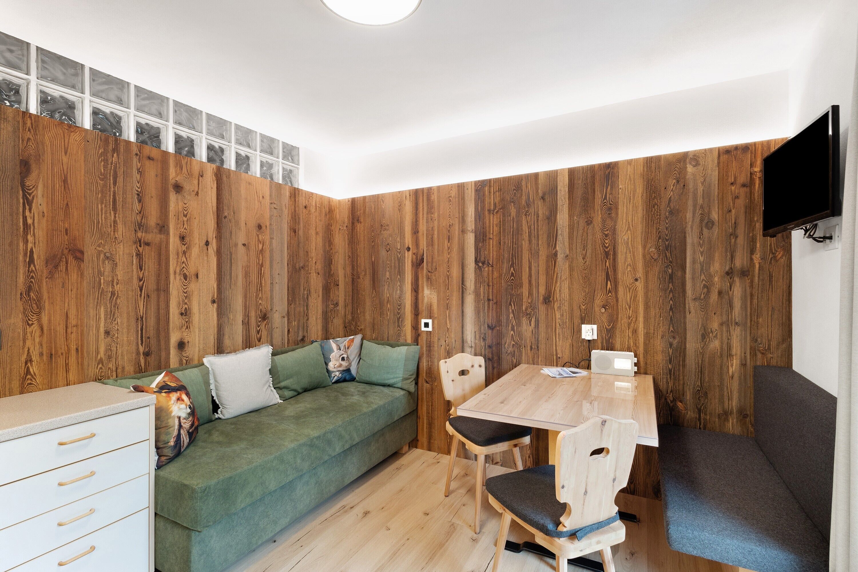 Ein Zimmer mit einer Holzwand, einem grünen Sofa, einem Holzesstisch und einem an der Wand montierten Fernseher.