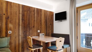 A room with a wooden wall panel, a TV mounted on the wall, a table with chairs, and a window with curtains. - Apartment 'Pfaffingerhof Apt Piccolo Rabbit' with Mountain View, Shared Garden and Wi-Fi (Anterselva di Sotto BZ)
