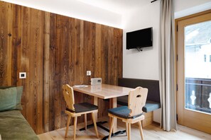 A room with a wooden wall panel, a TV mounted on the wall, a table with chairs, and a window with curtains. - Apartment 'Pfaffingerhof Apt Piccolo Rabbit' with Mountain View, Shared Garden and Wi-Fi (Anterselva di Sotto BZ)