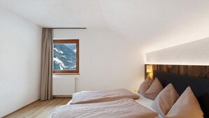 Een slaapkamer met een houten vloer, een bed met beige beddengoed, een raam met uitzicht op besneeuwde bergen en een wandlamp.