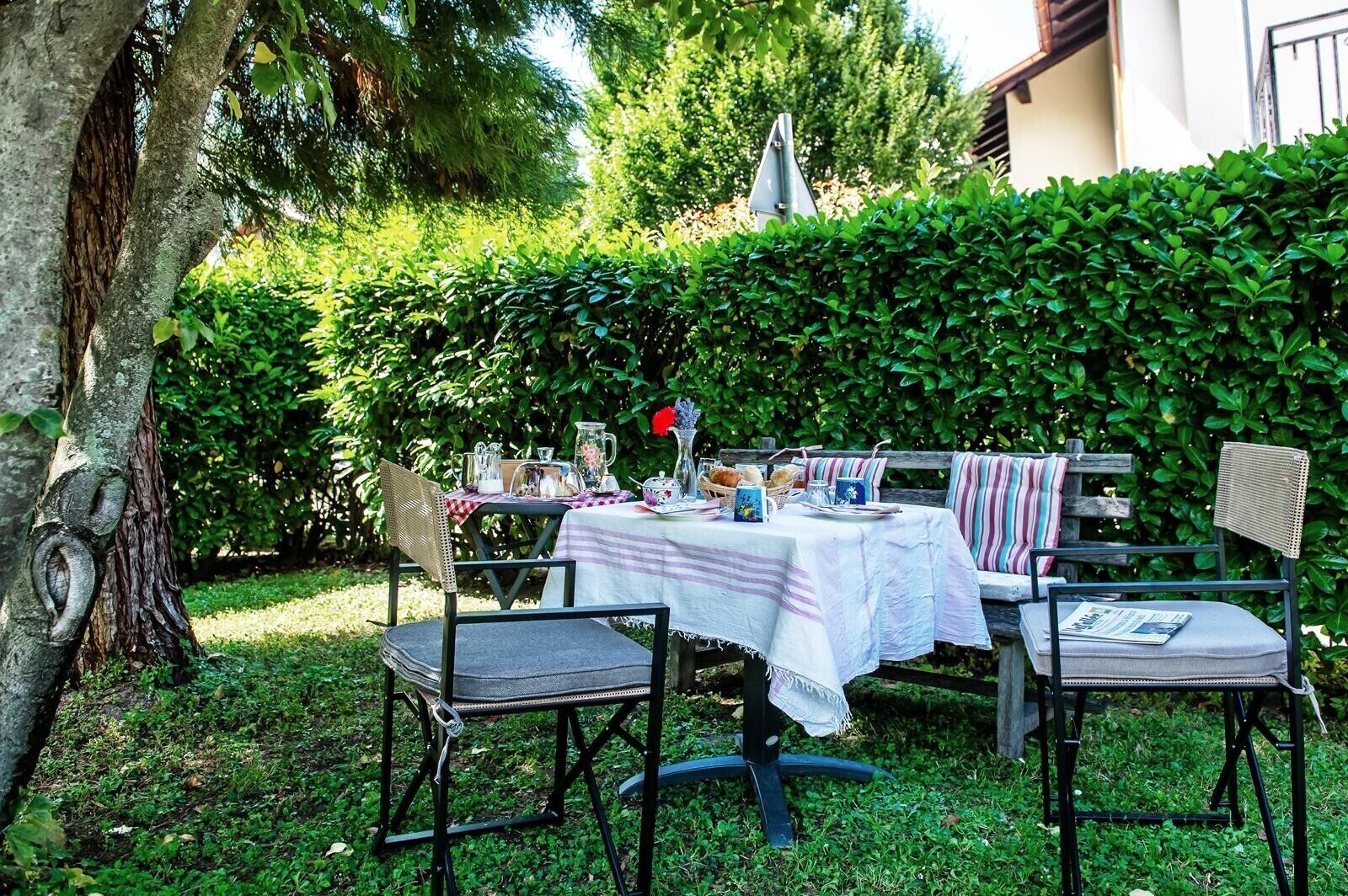 Area pranzo all'aperto con tavolo apparecchiato per due, circondata da vegetazione e un siepe.