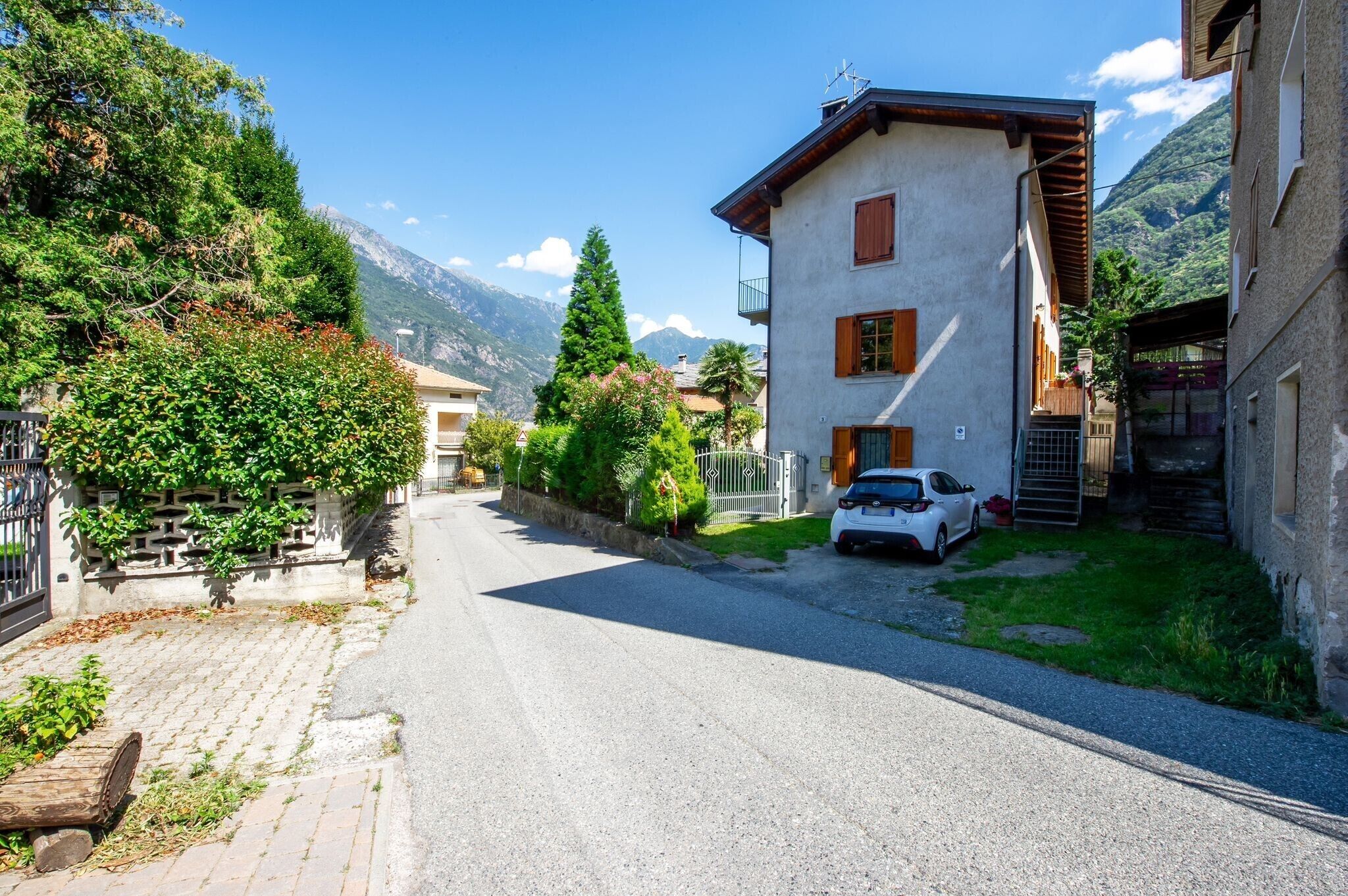 Una stretta via con un'auto bianca parcheggiata a destra, circondata da edifici con balconi e uno sfondo montuoso.
