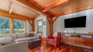 A spacious living room with a wooden ceiling, a large flat-screen TV, a sofa, a red patterned armchair, and a wooden cabinet. - Wapato Point Escape: Private Hot Tub • Walk to Beach • Lake Views (Manson)