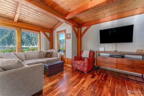 A spacious living room with a wooden ceiling, a large flat-screen TV, a sofa, a red patterned armchair, and a wooden cabinet.