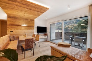 Een moderne woonkamer met een houten plafond, een flatscreen televisie, een eethoek en een balkon met uitzicht op een bebost gebied.