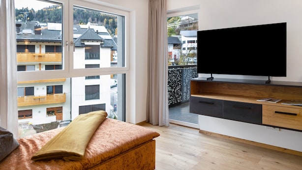 A modern living room with a large window, a flat-screen TV, and a wooden bench. - Apartment 'Paradeis Apartments A17' with Mountain View, Balcony and Wi-Fi (Mühlbach)