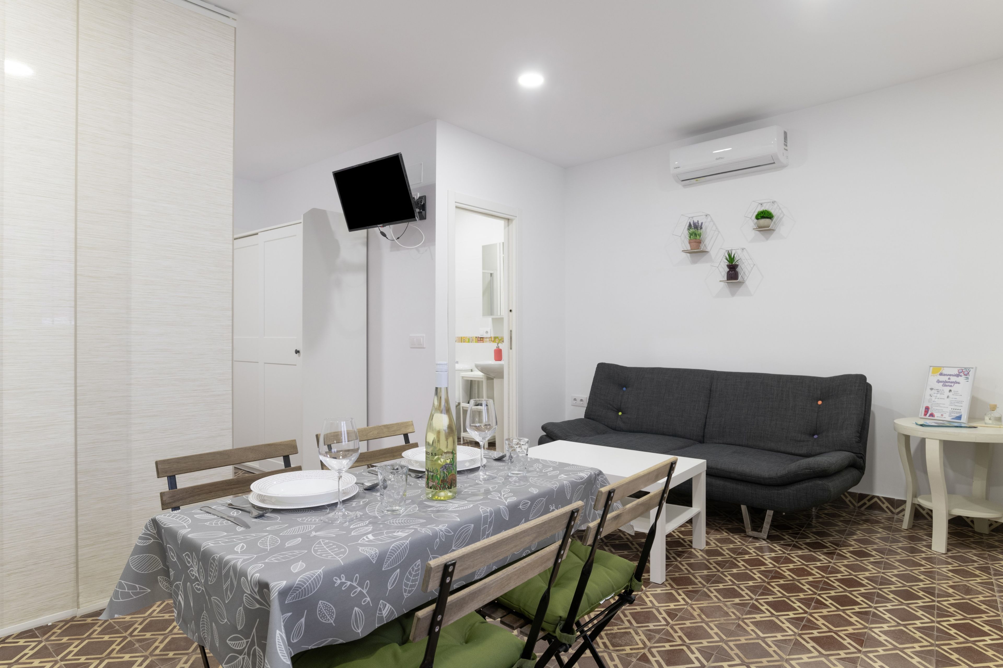 A dining area with a table set for two, a grey sofa, and a small side table with a sign.