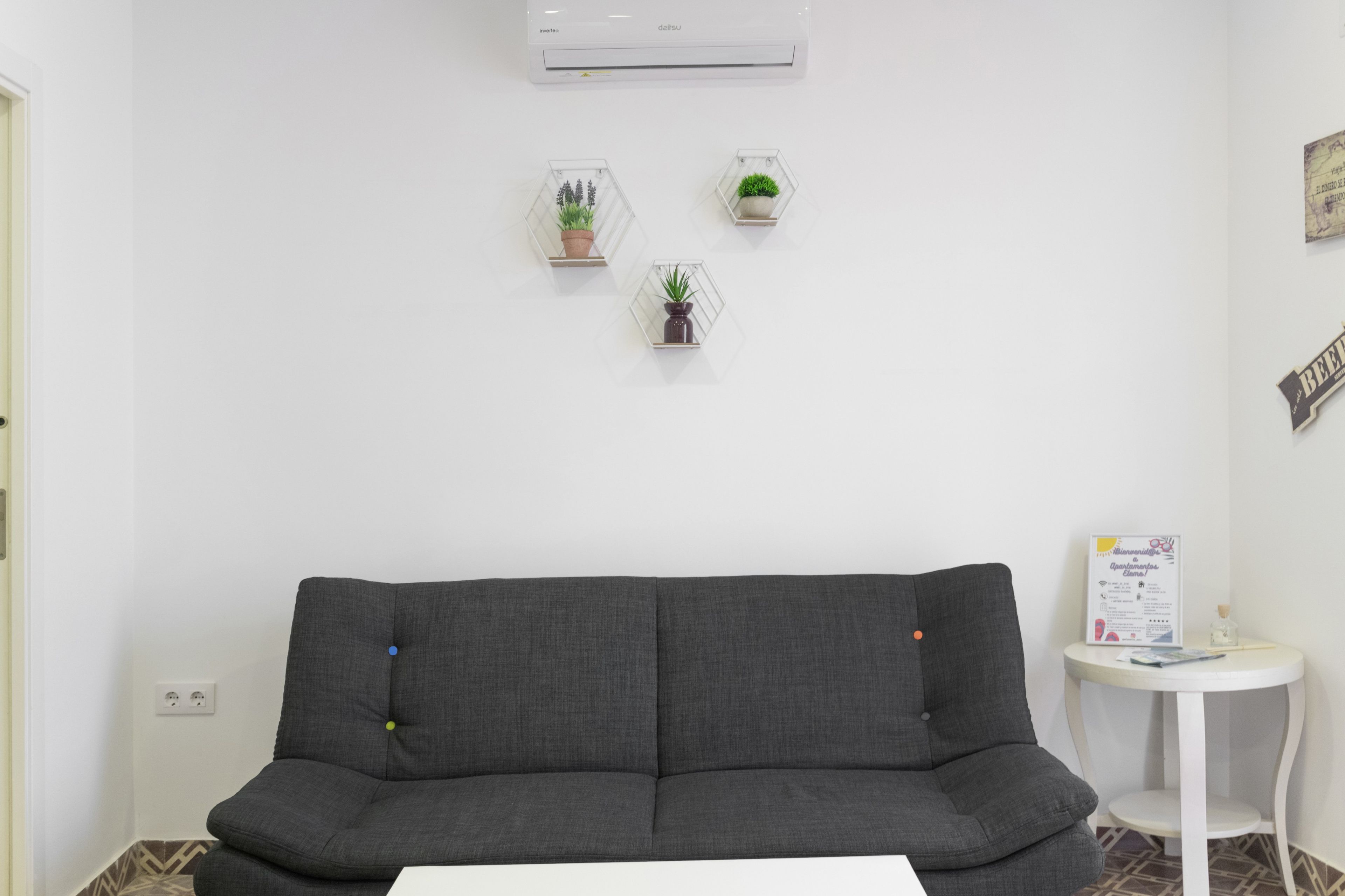 Un canapé aux couleurs neutres, une petite table blanche et trois plantes en pot disposées sur un mur blanc.