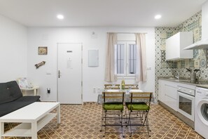 Een compact woongedeelte met een kitchenette, een eethoek met tafel en stoelen, een bank en een wasmachine.