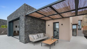 Un espacio exterior moderno con un muro de piedra, un sofá blanco, una mesa de madera y una puerta de cristal que da a una zona de cocina.