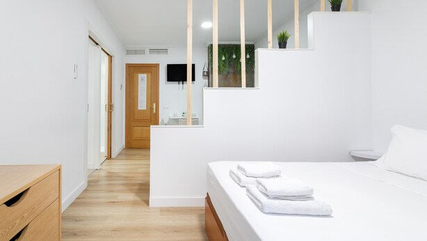 A modern bedroom with a wooden floor, a bed with white linens, a wooden dresser, and a view of a bathroom with a mirror and a plant.