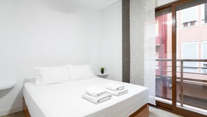 A hotel room with a bed, a small plant, a window with a view of buildings, and a sliding door.