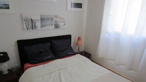 Een bed met een witte dekbedovertrek, twee donkere kussens, een nachtkastje met een lamp en ingelijste foto's aan de muur.