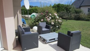 Een terras buiten met rieten meubilair, een witte schotelantenne en een tuin met bloemen en gras.