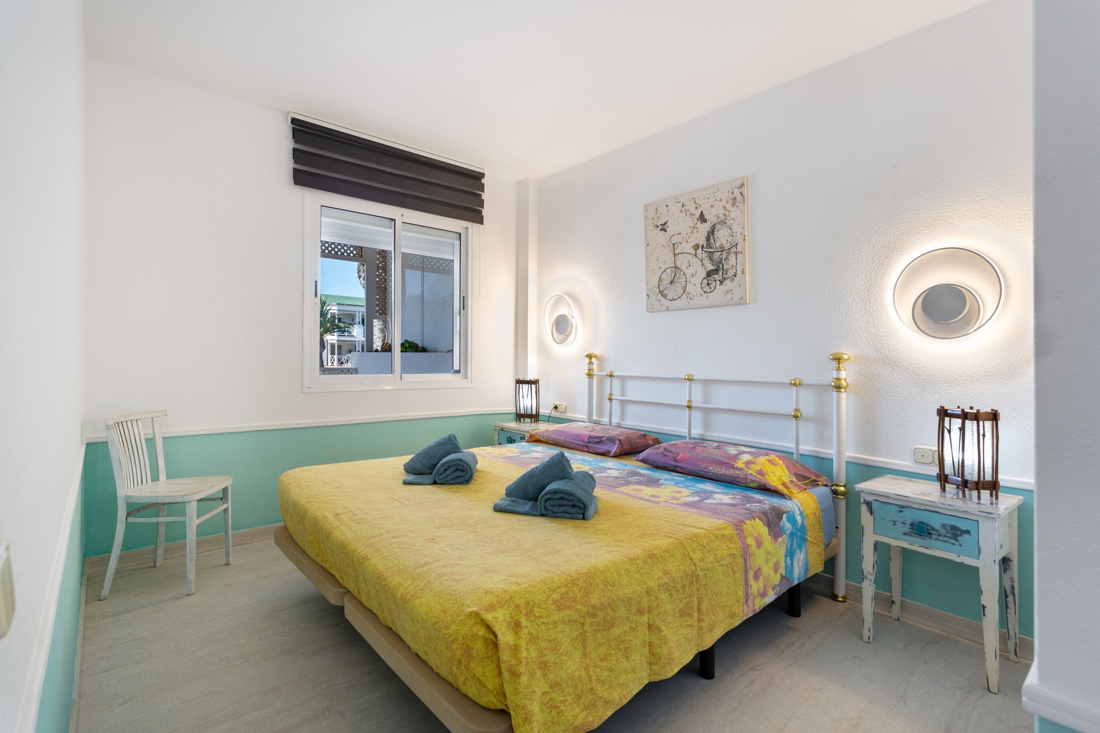 Un dormitorio con una cama, una silla, una mesita de noche, un ventanal y una lámpara fijada a la pared.