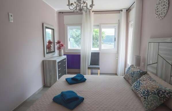A bedroom with a bed, a chair, a dresser, a window with curtains, and a view of greenery outside.