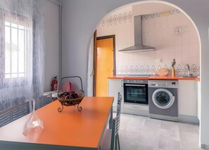A modern kitchen with an oven, dishwasher, and washing machine, a dining table with a fruit bowl, and a window with a view.