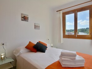 A bedroom with a bed, pillows, a window with a view, and a bedside table with a lamp. - Cosy Front-Line Beach Apartment in Mallorca (Son Serra de Marina)