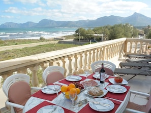 A dining table set with plates, cutlery, a bottle, and a fruit bowl, overlooking a coastal landscape with mountains in the distance. - Cosy Front-Line Beach Apartment in Mallorca (Son Serra de Marina)