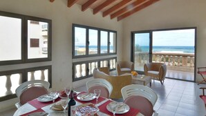 A dining area with a table set for two, large windows with ocean views, and a balcony with wicker furniture. - Cosy Front-Line Beach Apartment in Mallorca (Son Serra de Marina)