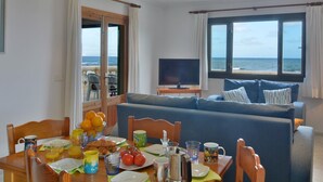 A room with a dining table set for breakfast, a sofa with cushions, a TV, and a view of the sea through large windows. - Cosy Front-Line Beach Apartment in Mallorca (Son Serra de Marina)