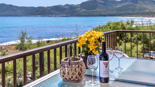 A table with a bottle of wine, glasses, and a basket of flowers on a balcony overlooking a coastal view. - Beautiful Front-Line Beach House in Mallorca (Son Serra de Marina)
