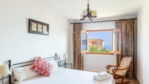A hotel room with a bed, a chair, a nightstand, a lamp, a picture frame, a window with a view, and a chandelier. - Beautiful Front-Line Beach House in Mallorca (Son Serra de Marina)