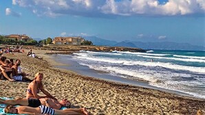 A beach with people sunbathing, a rocky shoreline, and buildings in the background. - Beautiful Front-Line Beach House in Mallorca (Son Serra de Marina)