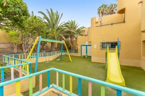 A playground with swings and a slide, surrounded by palm trees and a building with a yellow exterior.