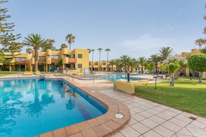 Une piscine avec une terrasse carrelée, des palmiers et un bâtiment doté de balcons.