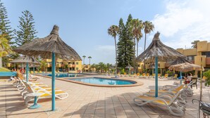 A pool area with sun loungers and thatched umbrellas, surrounded by palm trees and buildings.