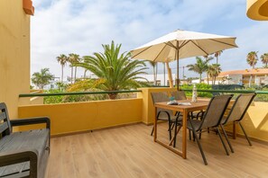 A balcony with a table and chairs, a large umbrella, and a view of palm trees and buildings.