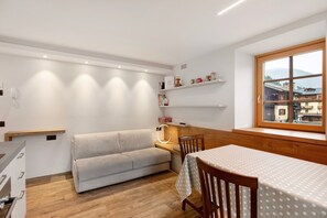 Un espacio habitable compacto con sofá, mesa de comedor, suelo de madera, estanterías empotradas y ventana con vistas.
