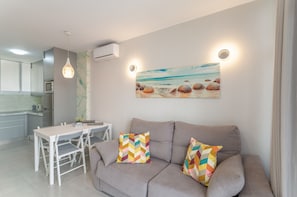 A modern living room with a grey sofa, a dining table, and chairs. There is a painting of a beach scene on the wall.
