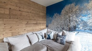 A living room with a wooden wall, a sofa with cushions, and a winter scene mural. - Apartment 'Le Chappart - Carroz D'Arâches' with Mountain View, Private Terrace and Wi-Fi (Arâches-la-Frasse)