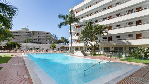A swimming pool with a tiled deck, surrounded by palm trees and a multi-story building with balconies.