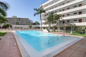 Una piscina con plataforma de baldosas, rodeada de palmeras y un edificio de varios pisos con balcones.