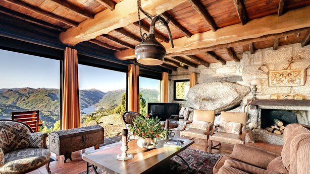 A cozy living room with a stone fireplace, wooden beams, and a large window overlooking a mountainous landscape. - Holiday Home 'Casa Do Gêres' with Lake View, Private Pool and Wi-Fi (Gêres)