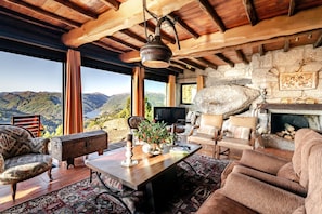 A cozy living room with a stone fireplace, wooden beams, and a large window overlooking a mountainous landscape. - Holiday Home 'Casa Do Gêres' with Lake View, Private Pool and Wi-Fi (Gêres)