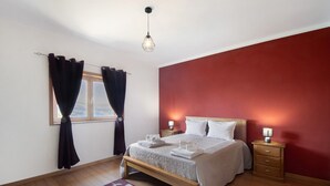 A bedroom with a bed, bedside tables, a nightstand, a window with curtains, and a hanging light fixture.