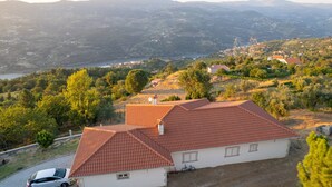 A house with a red-tiled roof, surrounded by trees and hills, overlooking a body of water.