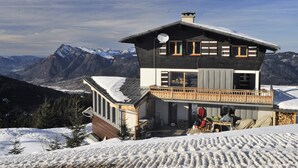 A two-story chalet with a balcony, snow-covered roof, and a view of mountains.