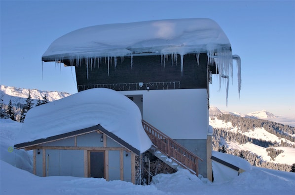 A snow-covered building with a wooden staircase leading up to a door, surrounded by a snowy landscape and mountains in the background.